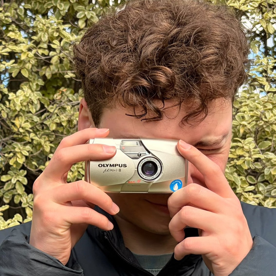 A young man taking a photo with the Olympus Mju II 35mm film camera in silver, a compact vintage camera