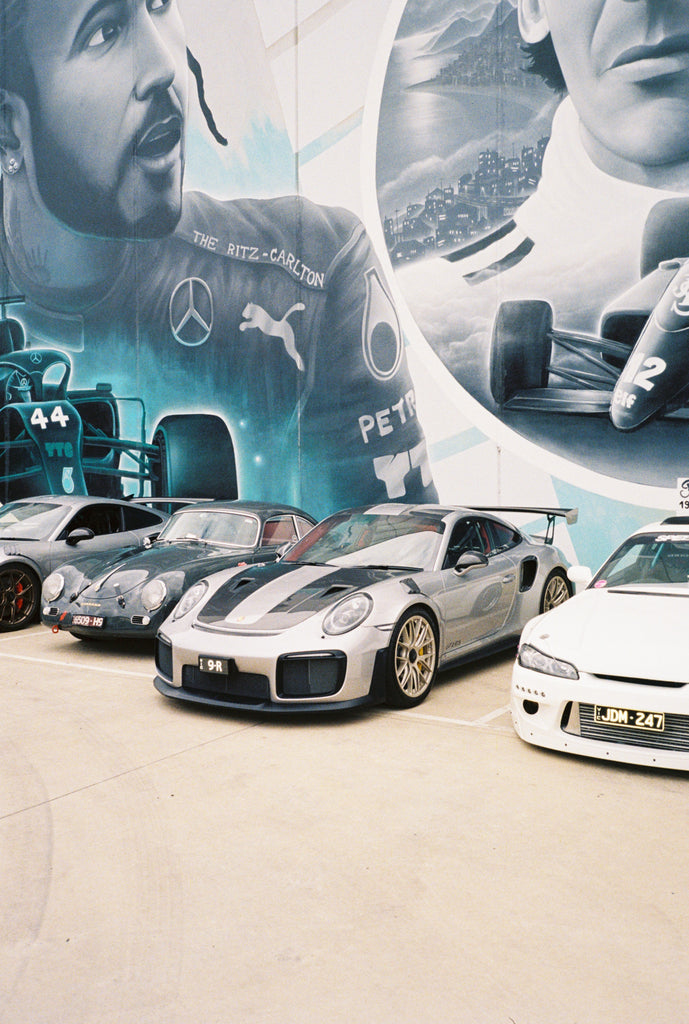Supercars and Japanese cars parked in-front of a Lewis Hamilton mural