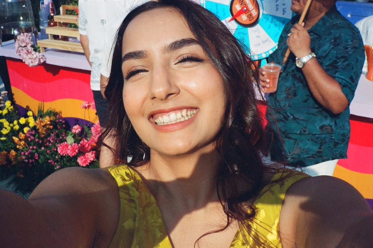 A sample phot taken with the Contax T2 (Gold) - A smiling woman taking a selfie outdoors, wearing a yellow sleeveless top. A man in a green patterned shirt stands in the background near colorful flowers and a decorated event setup.