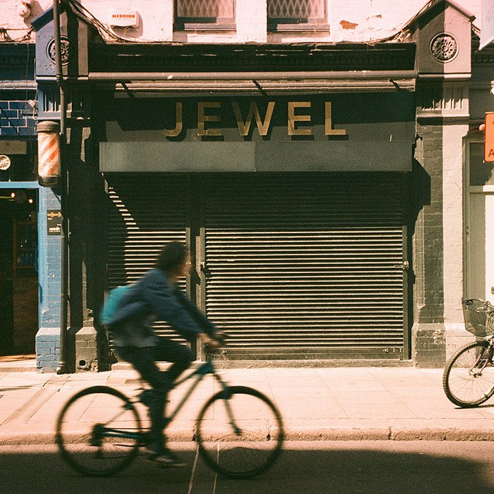 Sample photo taken with the Pentax Espio 160 35mm film camera. Man riding a bicycle past a closed jewelry store with 'Jewel' displayed on the shutter.